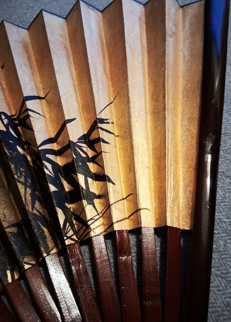 May include: A close-up of a traditional hand fan. The fan features a bamboo design in black ink on a gold and tan background. The ribs are a dark brown color.