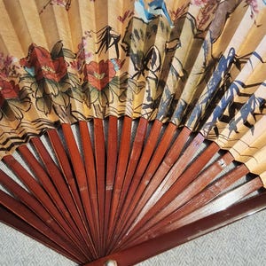 May include: A hand fan with a painted design featuring red flowers, a blue bird, and mountain scenery. The fan's paper is a light tan color, and the wooden ribs are a dark brown. A decorative item.