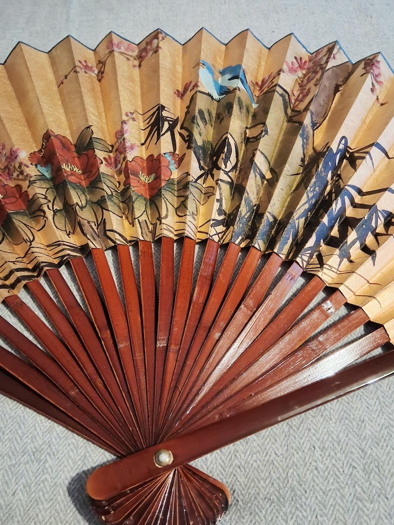 May include: A hand fan with a painted design featuring red flowers, a blue bird, and mountain scenery. The fan's paper is a light tan color, and the wooden ribs are a dark brown. A decorative item.
