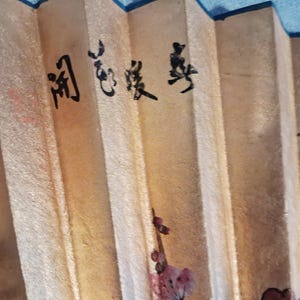 May include: A close-up of a beige folding fan with black calligraphy and floral accents. The fan has a textured surface and is partially open, revealing the pleated design. The calligraphy is in a vertical format.
