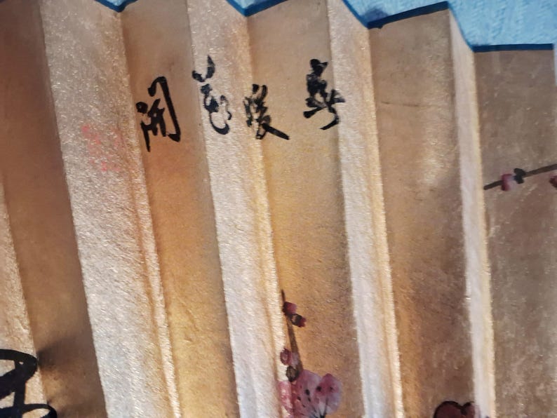 May include: A close-up of a beige folding fan with black calligraphy and floral accents. The fan has a textured surface and is partially open, revealing the pleated design. The calligraphy is in a vertical format.
