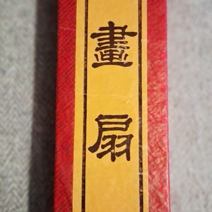 May include: A close-up of a red and yellow box with Chinese characters and the words "PAINTING FANS". The box has a textured red pattern and a yellow label with black text and a fan illustration.