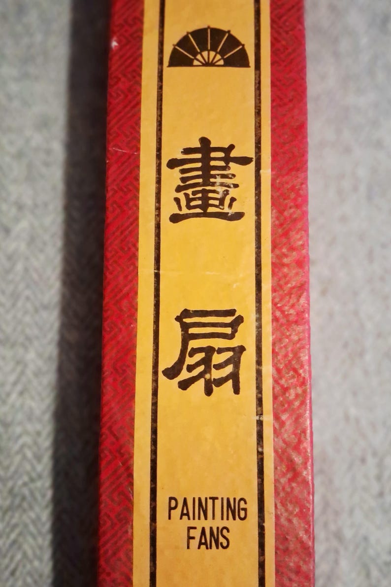 May include: A close-up of a red and yellow box with Chinese characters and the words "PAINTING FANS". The box has a textured red pattern and a yellow label with black text and a fan illustration.