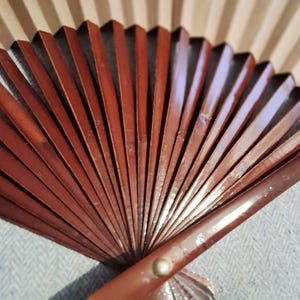 May include: A partially open, brown wooden hand fan with a decorative, fan-shaped base. The fan's ribs are a rich, reddish-brown color, and the background features a light beige, striped pattern. The fan is partially open, revealing its intricate design.