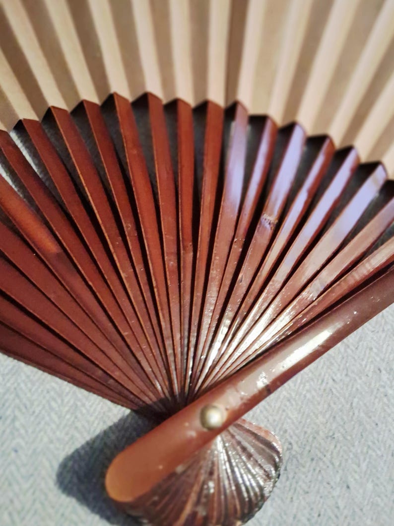 May include: A partially open, brown wooden hand fan with a decorative, fan-shaped base. The fan's ribs are a rich, reddish-brown color, and the background features a light beige, striped pattern. The fan is partially open, revealing its intricate design.