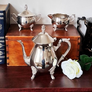 May include: A silver-plated tea set comprising a teapot with a lid, a lidded sugar bowl, and a creamer. Each piece features ornate, curved handles, decorative feet, and a polished, reflective surface. A white rose is placed beside the teapot.