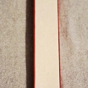 May include: A rectangular box with a red border and a cream-colored interior. The box is long and narrow, and it is sitting on a textured, gray surface.