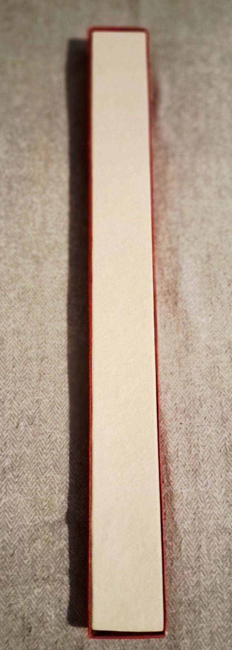 May include: A rectangular box with a red border and a cream-colored interior. The box is long and narrow, and it is sitting on a textured, gray surface.