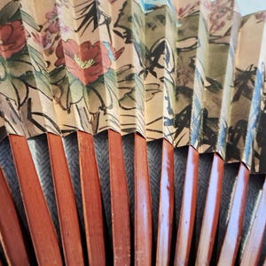 May include: A close-up of a decorative folding fan. The fan features a floral design with red and pink flowers, green leaves, and black outlines. The fan's wooden ribs are a reddish-brown color.
