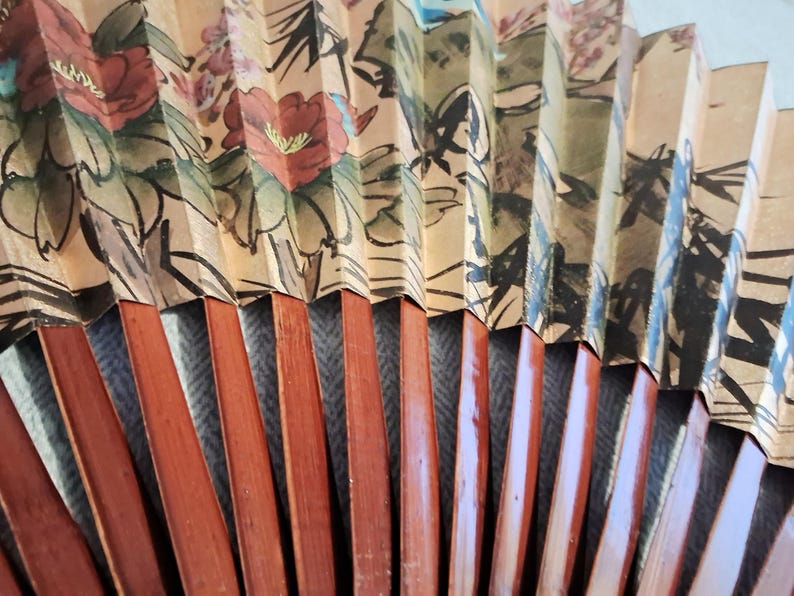 May include: A close-up of a decorative folding fan. The fan features a floral design with red and pink flowers, green leaves, and black outlines. The fan's wooden ribs are a reddish-brown color.