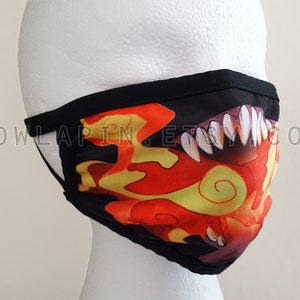 Puede incluir: Mascarilla negra con un diseño de llamas naranja y amarillo y dientes blancos afilados. La mascarilla tiene un borde negro y el texto "OWLAPIN.ETSY.COM" está impreso en el lateral.