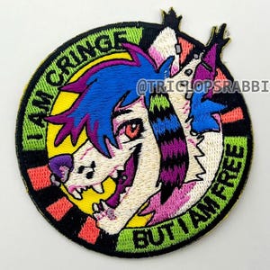 May include: Embroidered patch with a cartoon wolf-like character with blue hair and red eyes. The patch is circular, with the text "I AM CRINGE BUT I AM FREE" in a green and yellow border. The character's face is white, with purple, pink, and black accents.