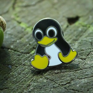 May include: A metal enamel pin featuring a cartoon penguin with a black body, white belly, and yellow beak and feet. The penguin has large white eyes with black pupils. A green acorn is positioned to the left of the penguin pin.