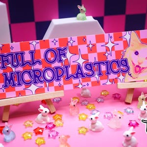 May include: A pink and orange sign on a wooden easel reads "FULL OF MICROPLASTICS" with a cartoon cat illustration. The image is surrounded by small bunny figurines and colorful star-shaped decorations.