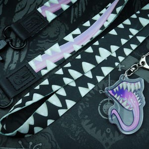 May include: A black fabric lanyard featuring a repeating pattern of white jagged teeth and a long pink tongue, finished with black metal hardware. An acrylic keychain displays a matching open mouth with sharp teeth and a long pink tongue.