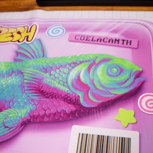 May include: A vibrant illustration of a coelacanth fish on a pink surface. The fish is rendered in blue, green, and pink hues, with the word "FRESH" in yellow and the word "COELACANTH" on a pink label. Decorative stickers are also present.