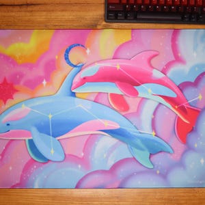 May include: A rectangular mousepad featuring a whimsical design of two dolphins in a dreamy, pastel-colored sky. One dolphin is blue with pink accents, the other is pink with red accents. A crescent moon and stars add to the celestial theme.
