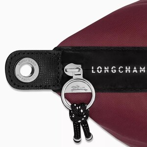May include: Close-up of a burgundy nylon bag with a black leather strap and silver zipper. The zipper pull features a silver ring with a horse logo and a black and white cord. The brand name "LONGCHAMP" is printed in white.