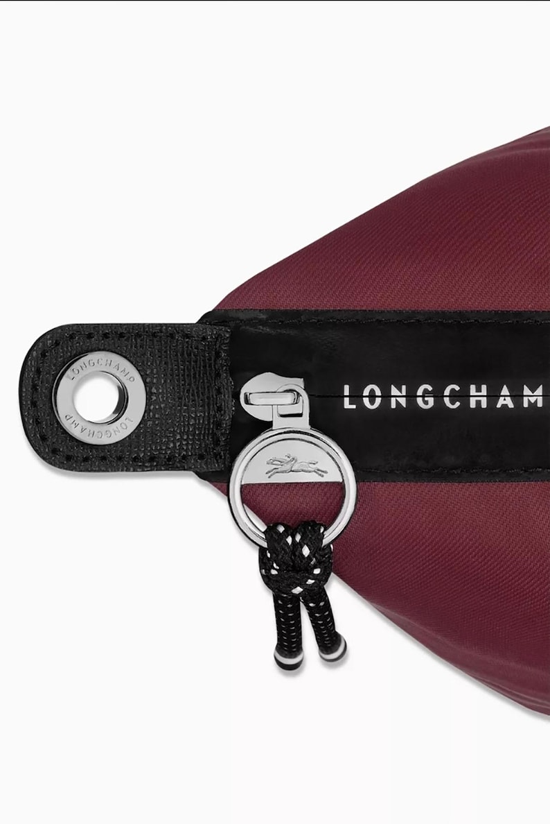 May include: Close-up of a burgundy nylon bag with a black leather strap and silver zipper. The zipper pull features a silver ring with a horse logo and a black and white cord. The brand name "LONGCHAMP" is printed in white.