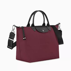 May include: A burgundy tote bag with black leather handles and a black shoulder strap. The bag has a flap closure and a small pocket on the front. The bag is made of a durable material and has silver-tone hardware.