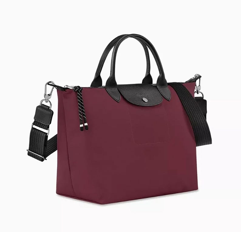 May include: A burgundy tote bag with black leather handles and a black shoulder strap. The bag has a flap closure and a small pocket on the front. The bag is made of a durable material and has silver-tone hardware.