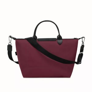May include: A burgundy tote bag with black leather handles and a black adjustable shoulder strap. The bag has a zippered top closure and silver-tone metal hardware. The bag is designed for everyday use.