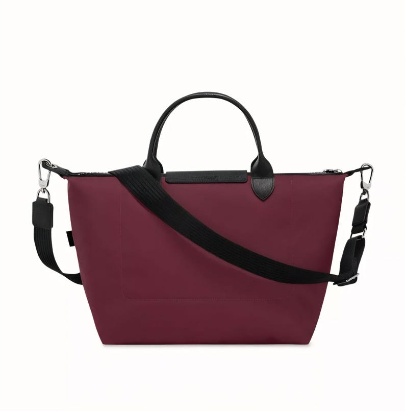 May include: A burgundy tote bag with black leather handles and a black adjustable shoulder strap. The bag has a zippered top closure and silver-tone metal hardware. The bag is designed for everyday use.