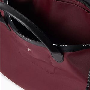 May include: A burgundy tote bag with black leather handles and trim. The interior is a matching burgundy color. The bag has a zippered closure and a small pocket on the front. The brand name is printed on the black trim.