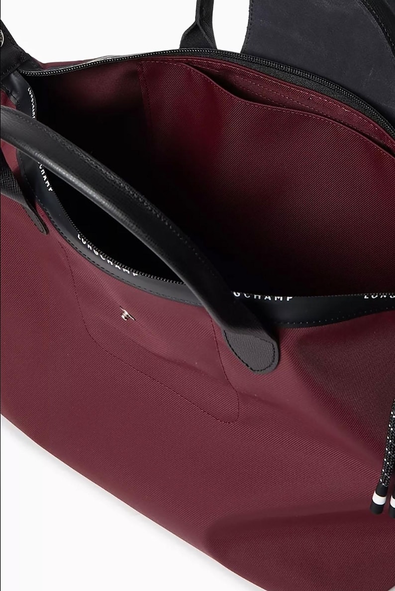 May include: A burgundy tote bag with black leather handles and trim. The interior is a matching burgundy color. The bag has a zippered closure and a small pocket on the front. The brand name is printed on the black trim.