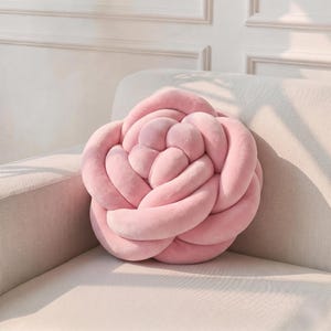 以下が含まれることがあります： 淡いピンク色の花の形をした装飾用クッション。クッションは柔らかく、豪華な素材で作られており、バラに似た結び目のデザインが特徴です。クッションは明るい色のソファの上に置かれ、居心地の良い魅力的な美学を作り出しています。