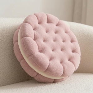 Puede incluir: Un cojín redondo y suave con forma de galleta sándwich rosa. La galleta tiene una superficie texturizada con pequeñas hendiduras espaciadas uniformemente. El relleno es de color crema, creando un diseño divertido y atractivo.