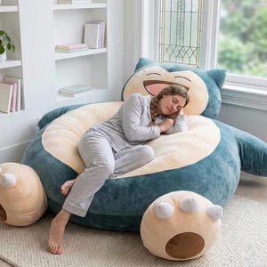 Può includere: Un grande pouf a forma di Snorlax, blu e crema. Il pouf ha una faccia sorridente ed è circondato da due cuscini a forma di piede abbinati. Una persona in pigiama a righe si riposa sul pouf.