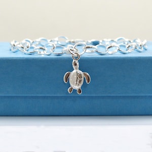 May include: A silver bracelet with a turtle charm. The bracelet features a chain link design and a small, detailed turtle pendant. The turtle charm has a textured shell and is suspended from the bracelet's chain. The bracelet is displayed on a blue surface.