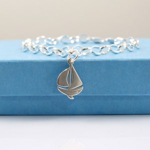 May include: A silver charm bracelet featuring a sailboat pendant. The bracelet has a chain link design, and the sailboat charm has a mast, sail, and hull. The bracelet is displayed on a blue box.