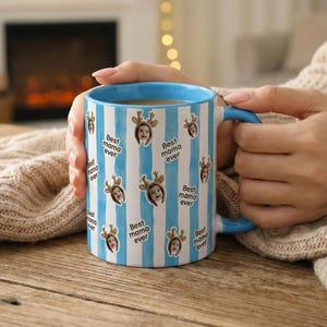 May include: A blue and white striped ceramic mug with a blue handle and rim. The mug is decorated with images of a child wearing reindeer antlers and the text "Best mama ever".
