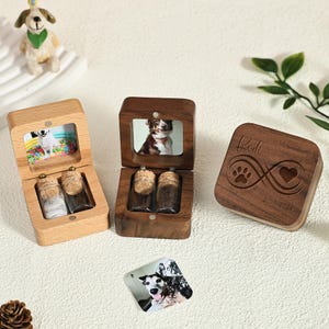 May include: Pet memorial boxes made of wood, featuring a photo frame and small glass bottles. One box is open, showing a dog photo and two bottles with cork stoppers. The other box is closed, with a paw print and infinity symbol.