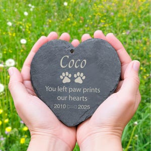 Może przedstawiać: Szary, w kształcie serca kamień pamiątkowy z łupków z wygrawerowanym imieniem "Coco" i odciskami łap. Napis głosi "You left paw prints our hearts 2010 2025". Kamień trzymany jest w dłoniach na rozmytym zielonym tle.