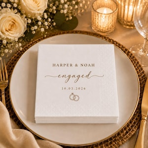May include: A stack of white square paper napkins featuring a textured border and gold-coloured text that reads HARPER & NOAH, engaged, and 10.03.2026, accompanied by a small graphic of two interlocking rings positioned below the date.