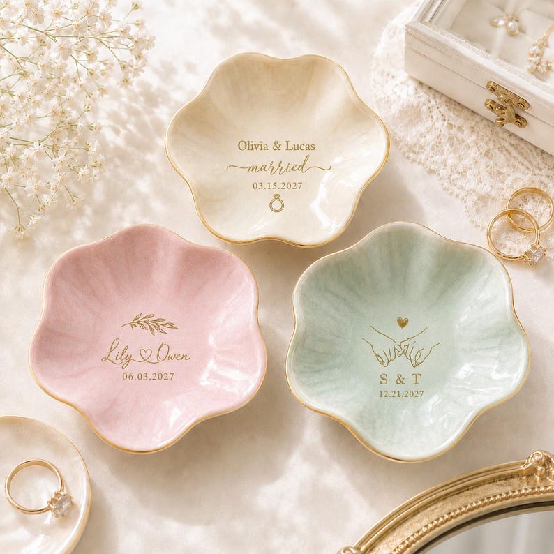 K&ouml;nnte beinhalten: Drei blumenf&ouml;rmige Schmuckschalen aus Keramik in Creme, zartem Rosa und Hellgr&uuml;n mit goldfarbenen R&auml;ndern. Jede Schale ist mit individuellen goldenen Schriftz&uuml;gen und Grafiken versehen, darunter Namen, Daten sowie Ring-, Herz- und Handsymbole.