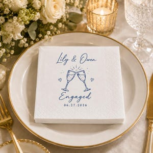 May include: A square white paper napkin with a textured border featuring blue printed text that reads Lily & Owen Engaged 06.27.2026, accompanied by a line art illustration of two clinking champagne flutes and two small hearts.