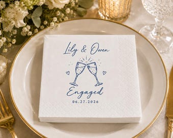 May include: A square white paper napkin with a textured border featuring blue printed text that reads Lily & Owen Engaged 06.27.2026, accompanied by a line art illustration of two clinking champagne flutes and two small hearts.