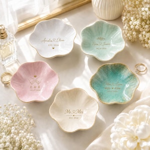 K&ouml;nnte beinhalten: F&uuml;nf blumenf&ouml;rmige Schmuckschalen aus Keramik in Wei&szlig;, Mintgr&uuml;n, zartem Rosa, T&uuml;rkis und Creme. Jede Schale ist mit goldfarbenen, personalisierten Texten versehen, darunter Namen wie Amelia & Oliver, Nora & James sowie verschiedene Daten.