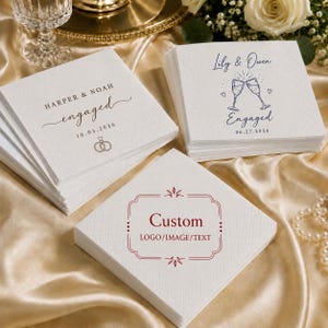 May include: Three stacks of square white paper napkins with embossed borders. One stack features the text HARPER & NOAH engaged 10.03.2026 with ring icons. Another shows Lily & Owen Engaged 06.27.2026 with champagne flutes. The third displays Custom LOGO/IMAGE/TEXT.
