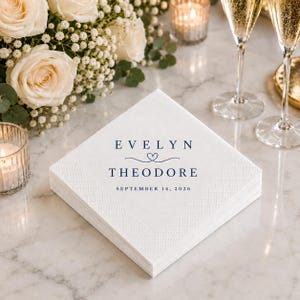 May include: A square white paper napkin with a textured border, featuring dark blue printed text that reads EVELYN, a small heart icon, THEODORE, and the date SEPTEMBER 14, 2026, centred on the front surface of the folded stack of napkins.