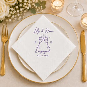 May include: A white square paper napkin featuring a purple ink print of two clinking champagne glasses, small hearts, and the text Lily & Owen Engaged 06.27.2026. The napkin is centred on a white plate with a gold rim, flanked by a gold fork and knife.