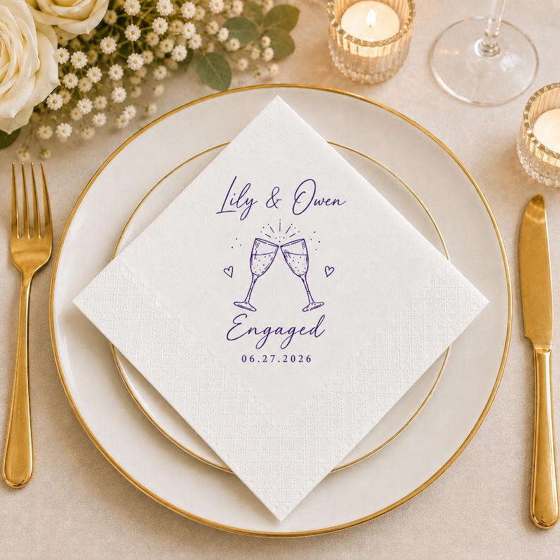 May include: A white square paper napkin featuring a purple ink print of two clinking champagne glasses, small hearts, and the text Lily & Owen Engaged 06.27.2026. The napkin is centred on a white plate with a gold rim, flanked by a gold fork and knife.