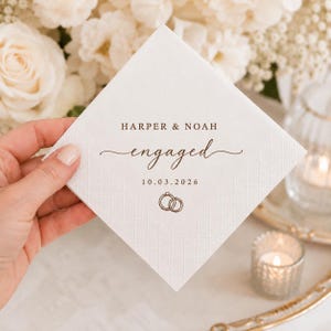 May include: A square white paper napkin with a textured diamond border pattern. The centre features brown printed text reading HARPER & NOAH, the word engaged in a script font, the date 10.03.2026, and a small icon of two interlocking rings.