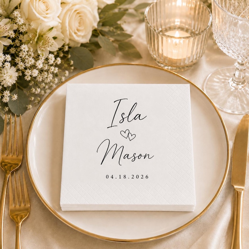May include: A square white paper napkin featuring black printed text that reads Isla, followed by two small heart icons, the name Mason, and the date 18.04.2026. The napkin has a subtle embossed diamond pattern along its border and sits on a plate.