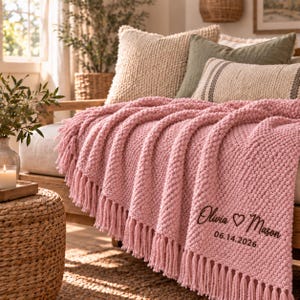 May include: A soft pink knitted throw blanket with a textured bobble pattern and long fringe trim. The blanket features black embroidered text that reads Olivia heart Mason and the date 14.06.2026, draped over a sofa with various pillows.