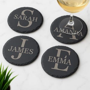 Può includere: Quattro sottobicchieri rotondi in ardesia grigio scuro con testo personalizzato. Ogni sottobicchiere presenta un nome: Sarah, Amanda, James ed Emma. Un bicchiere di vino bianco è sopra uno.